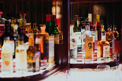 A well-lit bar shelf displaying an assorted collection of liquor bottles, including whiskey, vodka, and other spirits, organized neatly with some glassware placed in front. The atmosphere suggests a cozy, inviting setting.
