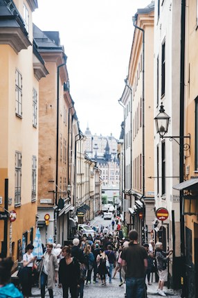A bustling narrow street lined with historic buildings, filled with people walking and engaging with shops and cafes. The architecture features warm-toned facades and a view of an ornate structure in the distance.