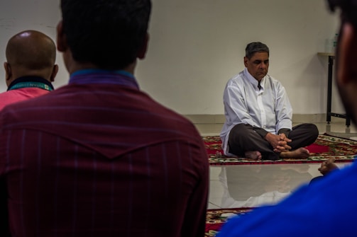 A man in a white shirt sits cross-legged on a colorful rug with his hands clasped, possibly in meditation or deep thought, while several people sit facing him with their backs to the camera. The room has a calm atmosphere and is simply furnished.