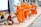 An elderly woman sits on a sidewalk with various baskets in front of her, offering food to a line of young monks dressed in bright orange robes. Each monk is holding a bowl, receiving alms. Two dogs are sitting and standing nearby, observing the scene. The setting appears to be a street with some buildings in the background.