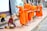 An elderly woman sits on a sidewalk with various baskets in front of her, offering food to a line of young monks dressed in bright orange robes. Each monk is holding a bowl, receiving alms. Two dogs are sitting and standing nearby, observing the scene. The setting appears to be a street with some buildings in the background.