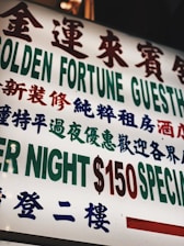 A brightly lit sign with text in both Chinese and English, featuring large, bold characters advertising a guesthouse. The sign prominently displays pricing information in red and green.
