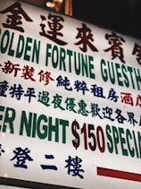 A brightly lit sign with text in both Chinese and English, featuring large, bold characters advertising a guesthouse. The sign prominently displays pricing information in red and green.