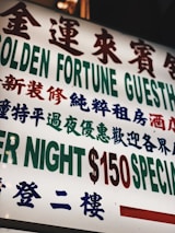 A brightly lit sign with text in both Chinese and English, featuring large, bold characters advertising a guesthouse. The sign prominently displays pricing information in red and green.