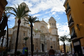 A historic cathedral with a large, ornate facade and tall towers under a partly cloudy sky. Palm trees flank the building, and a sunbeam glints off one of the towers. People are gathered in the square in front, surrounded by quaint buildings.
