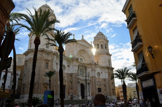A historic cathedral with a large, ornate facade and tall towers under a partly cloudy sky. Palm trees flank the building, and a sunbeam glints off one of the towers. People are gathered in the square in front, surrounded by quaint buildings.