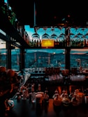 A bar interior with a cityscape view through large windows, showcasing bottles and glassware. Hanging wine glasses and a sign that reads 'Eleven BCN World Class' are visible. The scene is dimly lit with a mix of moody blues and warm tones.