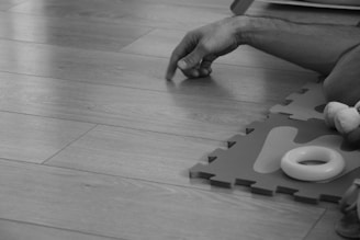 A black and white image shows a hand reaching out on a wooden floor next to a puzzle mat. The hand appears relaxed and there is a toy ring and part of a plush toy visible.