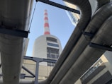 Large industrial pipes frame a view of a tall chimney with red and white stripes against a clear blue sky. The structure is made of concrete and metal, typical of a power plant or industrial facility.