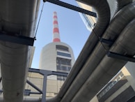 Large industrial pipes frame a view of a tall chimney with red and white stripes against a clear blue sky. The structure is made of concrete and metal, typical of a power plant or industrial facility.