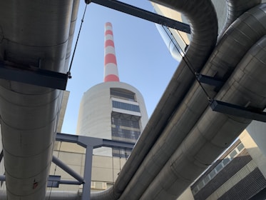 Large industrial pipes frame a view of a tall chimney with red and white stripes against a clear blue sky. The structure is made of concrete and metal, typical of a power plant or industrial facility.