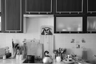 A well-organized kitchen countertop featuring various utensils and appliances. Wooden cabinets occupy the upper part of the image, while several cutting boards are lined up against the tiled wall. A kettle sits on the stovetop alongside other cooking utensils. A mug and a small pot are next to stacked plates and a grater. The kitchen has a clean and tidy appearance.