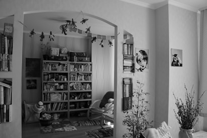 A cozy room filled with bookshelves, a variety of books, and assorted decorations. There are small plants, wall art featuring illustrations of faces, and a string of hanging stars and toy decorations. A wicker basket filled with textiles and a woven rug add a touch of homeliness to the space, while decorative suitcases sit atop the bookshelves.