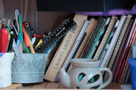 A cluttered shelf containing several notebooks, a ruler, and an assortment of pens and pencils in a metal container. Books are stacked horizontally against the notebooks, and a ceramic mug with cut-out patterns is in the foreground.