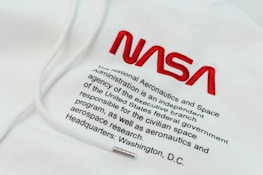 The image features a piece of clothing, likely a hoodie, with the NASA logo prominently embroidered in red. Below the logo, there is text that provides information about NASA, stating it as an independent agency of the United States federal government responsible for the civilian space program and aerospace research, with its headquarters in Washington, D.C.