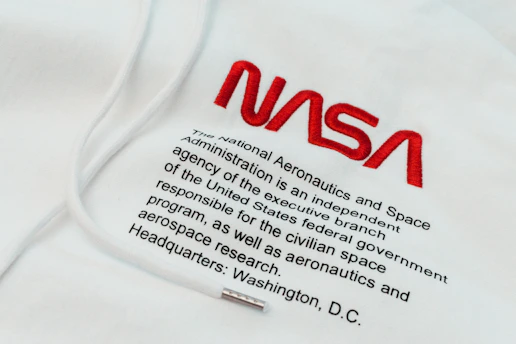 The image features a piece of clothing, likely a hoodie, with the NASA logo prominently embroidered in red. Below the logo, there is text that provides information about NASA, stating it as an independent agency of the United States federal government responsible for the civilian space program and aerospace research, with its headquarters in Washington, D.C.