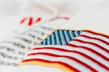 An embroidered American flag patch with vivid red and white stripes and blue stars on a white fabric background. Some out-of-focus text appears behind, adding depth to the image.