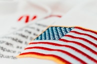 An embroidered American flag patch with vivid red and white stripes and blue stars on a white fabric background. Some out-of-focus text appears behind, adding depth to the image.