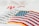 An embroidered American flag patch with vivid red and white stripes and blue stars on a white fabric background. Some out-of-focus text appears behind, adding depth to the image.