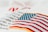 An embroidered American flag patch with vivid red and white stripes and blue stars on a white fabric background. Some out-of-focus text appears behind, adding depth to the image.
