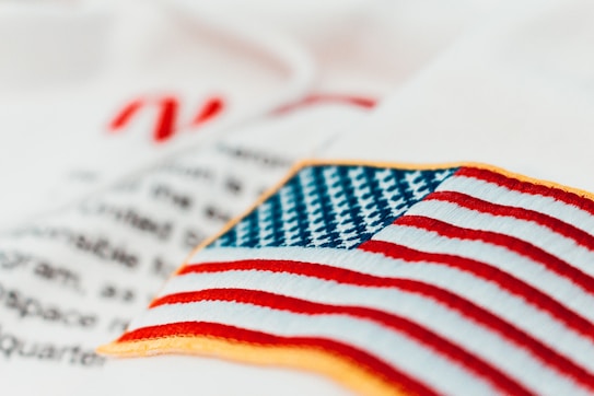 An embroidered American flag patch with vivid red and white stripes and blue stars on a white fabric background. Some out-of-focus text appears behind, adding depth to the image.
