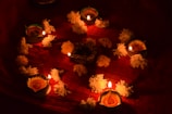 Lit oil lamps are arranged in a circular pattern on a red cloth, surrounded by white flowers. Each lamp emits a warm glow, creating an ambient and festive atmosphere. The candles, decorated with ornate designs, enhance the cultural and traditional vibe.