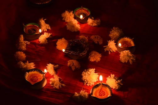 Lit oil lamps are arranged in a circular pattern on a red cloth, surrounded by white flowers. Each lamp emits a warm glow, creating an ambient and festive atmosphere. The candles, decorated with ornate designs, enhance the cultural and traditional vibe.