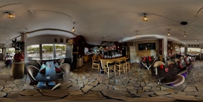 A spacious and modern restaurant interior featuring stone flooring and several dining tables with curved chairs. There is a bar with tall wooden stools and a variety of decor elements, including hanging lights and ornamental plants. Large windows allow natural light to enter, illuminating the space.