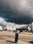 Dark storm clouds loom over a suburban area with buildings and parked cars. A person wearing a long garment walks along the street. Trees line the area, and a bank sign is visible, hinting at a commercial zone.