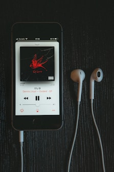 A smartphone displays a music player app with the song 'Gfy' by Dennis Lloyd from the 'Exident - EP' album. The album cover shows a red bird on a black background. White earphones are placed next to the phone on a dark wooden surface.