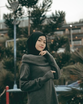 A person wearing a dark hijab and a knitted sweater stands in an outdoor setting with trees and a modern building in the background. The focus is on the person, who is posing with a thoughtful expression.
