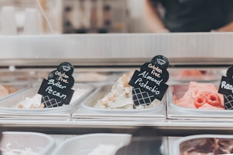 A display of different gluten-free ice cream flavors is shown in metal containers, each labeled with a black sign. Flavors include Butter Pecan, Almond Pistachio, and Raspberry. Toppings like nuts and fresh raspberries are visible on the ice cream.