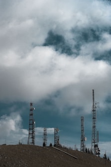 Multiple telecommunications towers rise against a backdrop of a cloudy sky. The towers are tall and metal, equipped with various antennas and satellite dishes. The scene suggests a rural or remote setting with a focus on the technology infrastructure.