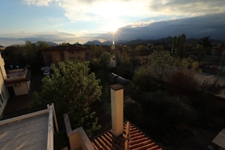 A scenic view of a residential neighborhood with several houses surrounded by trees. The sun is setting behind distant mountains, casting a warm glow over the landscape. The rooftops and a chimney are visible in the foreground. The sky is partly cloudy, adding texture to the scene.