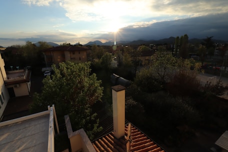 A scenic view of a residential neighborhood with several houses surrounded by trees. The sun is setting behind distant mountains, casting a warm glow over the landscape. The rooftops and a chimney are visible in the foreground. The sky is partly cloudy, adding texture to the scene.