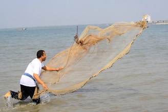 A person wearing a white shirt is casting a large fishing net into the water. The action is dynamic, with the net unfurling above the water surface, creating a sense of motion. The background features a calm sea and a distant shoreline with a boat.