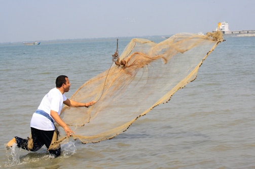 A person wearing a white shirt is casting a large fishing net into the water. The action is dynamic, with the net unfurling above the water surface, creating a sense of motion. The background features a calm sea and a distant shoreline with a boat.