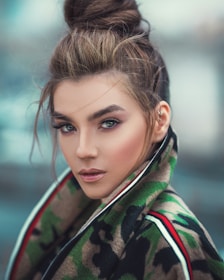 A person with an intricate updo hairstyle gazes intently at the viewer, wearing a camouflage-patterned jacket with red and white accents. The makeup is striking, with bold eye makeup highlighting the eyes.