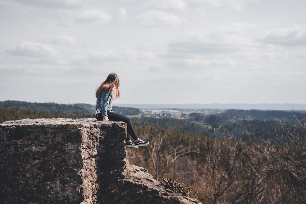 woman sitting on cliff's edge overlooking forest during daytime