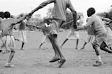 Children are holding hands and joyfully playing in a circle outdoors. They appear to be in school uniforms, enjoying a game in a natural setting with trees and open space around them.