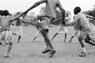 Children are holding hands and joyfully playing in a circle outdoors. They appear to be in school uniforms, enjoying a game in a natural setting with trees and open space around them.