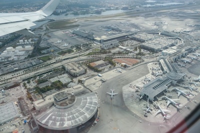 An aerial view of a large airport with multiple runways, terminals, and aircraft. Several planes are parked near the terminal gates, and taxiways connect the various parts of the airport. The background features a body of water and an urban landscape.