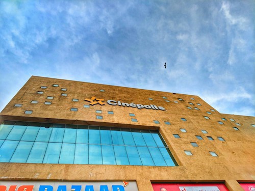 A modern building facade with a minimalist design featuring multiple small square windows scattered across a tan-colored wall. The structure is topped with a prominent sign displaying the name 'Cin&eacute;polis' in bold letters. The sky is clear with a single bird flying overhead, adding a dynamic element to the scene.