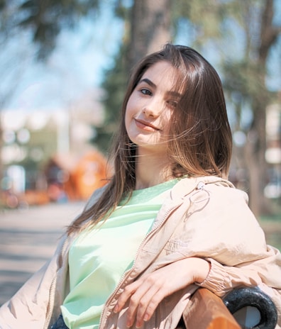 A young woman in a fitted casual blazer and jeans, sitting relaxed on a bench in a park.