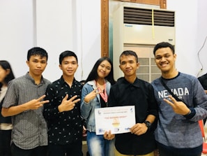 A group of five young adults stand together indoors, smiling and displaying a certificate. Two of them make peace signs with their hands. The person in the center holds the certificate. An air conditioning unit and a partially visible person are in the background.