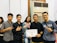 A group of five young adults stand together indoors, smiling and displaying a certificate. Two of them make peace signs with their hands. The person in the center holds the certificate. An air conditioning unit and a partially visible person are in the background.