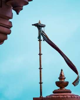 A tall, ornate flagpole with a golden, metallic finish is topped with a decorative element. A red and gold flag is attached, fluttering against a clear blue sky. An architectural structure with elaborate carvings is partially visible, showing traditional design elements.