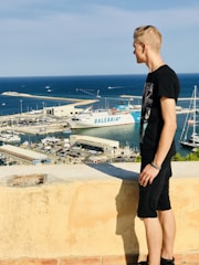 A man stands on a stone ledge overlooking a busy marina. In the harbor below, several boats and a large ship with 'Balearia' written on the side are docked. The sky is clear and the sea is a deep blue, with small waves visible. The man is wearing a black shirt and shorts and is looking towards the water.