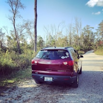 A red Chevrolet SUV is parked on a gravel path surrounded by tall trees and greenery. The vehicle has several stickers on its rear window, suggesting it may be used for travel or outdoor adventures. A clear blue sky is visible above, adding to the serene atmosphere of the location.