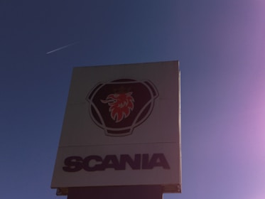 A tall sign displays a logo featuring a red griffin's head within a shield-like emblem, above the prominent text 'SCANIA'. The background is a clear blue sky with a faint jet contrail visible.